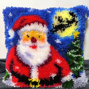 Puede incluir: Un coj&iacute;n cuadrado de ganchillo con un dise&ntilde;o texturizado de Santa Claus con traje rojo y barba blanca, de pie junto a un &aacute;rbol de Navidad verde sobre un cielo nocturno azul oscuro con una gran luna amarilla y detalles de nieve blanca.