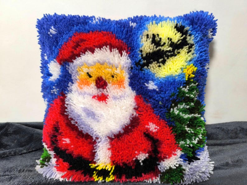 Puede incluir: Un coj&iacute;n cuadrado de ganchillo con un dise&ntilde;o texturizado de Santa Claus con traje rojo y barba blanca, de pie junto a un &aacute;rbol de Navidad verde sobre un cielo nocturno azul oscuro con una gran luna amarilla y detalles de nieve blanca.
