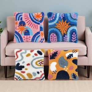 May include: Four square throw pillows with colorful abstract designs. The pillows feature a variety of patterns in shades of pink, blue, orange, yellow, and white. They are placed on a pink sofa.