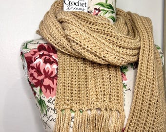 May include: A tan crocheted scarf with a ribbed texture and long fringe ends. The scarf is draped over a mannequin torso featuring a floral pattern and a white tag with the text Crochet Dreams and a yarn ball icon with two small stars.