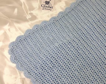 May include: A light blue crocheted blanket featuring a textured stitch pattern and a scalloped edge border. A small square white tag is attached to the blanket, displaying the text Crochet Dreams with a graphic of a ball of yarn.