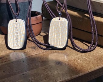 Puede incluir: Tres colgantes rectangulares con cordones marrón oscuro. Los colgantes tienen un fondo color crema con un borde negro. Un colgante tiene caracteres chinos, mientras que los otros tienen un patrón de cuadrícula. Los cordones tienen una cuenta plateada.