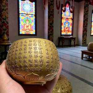Puede incluir: Un cuenco esférico dorado con texto en relieve. El cuenco tiene una base de flor de loto y se sostiene en una mano. El fondo presenta vidrieras y un interior de templo. La superficie del cuenco está cubierta de intrincados caracteres.