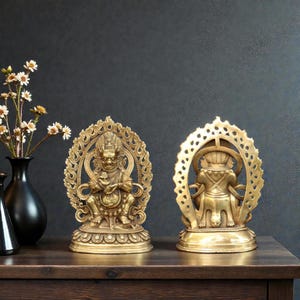 May include: Two brass-colored statues of seated figures with ornate halos. The front-facing statue has a detailed face and is holding an object. The other statue is a rear view. They are on a dark wooden surface.