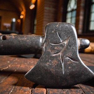 May include: Close-up of a dark, weathered axe head with a leaf design carved into the blade. The axe has a dark handle and is resting on a wooden surface. The axe appears to be made of metal.