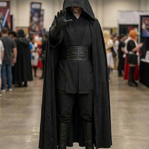Puede incluir: Una persona con un disfraz negro con capucha, cinturón y guantes negros, posando con la mano extendida. El disfraz incluye una capa larga, pantalones negros y botas. El fondo muestra una convención.
