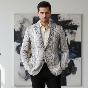 Peut inclure: Veste à motif peau de serpent gris et blanc, avec une fermeture à deux boutons. La veste est portée sur une chemise crème et un pantalon noir. L'arrière-plan présente de l'art abstrait.