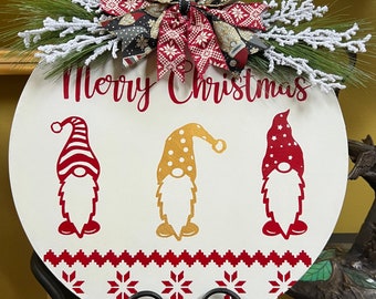May include: A white round wooden sign with the text "Merry Christmas" in red lettering. Three gnomes are depicted on the sign, one with a red hat, one with a gold hat, and one with a yellow hat. The sign has a red and white Scandinavian pattern at the bottom and is decorated with a red and black bow and white frosted pine sprigs.