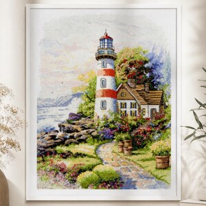 Könnte beinhalten: Ein gerahmtes Kreuzstichbild einer Küstenlandschaft mit einem rot-weiß gestreiften Leuchtturm, einem kleinen Haus und einem Steinweg. Das Kunstwerk ist von bunten Blumen und Grün umgeben, mit Blick auf das Meer und die Berge im Hintergrund.