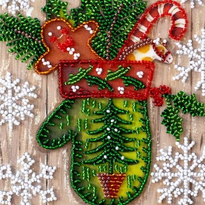 Könnte beinhalten: Ein bestickter Weihnachts-Fäustling mit Lebkuchenmann, Zuckerstange und Pferd. Der grüne Fäustling hat einen Weihnachtsbaum und einen Topf. Weiße Schneeflocken und ein roter Rand mit weißen Punkten vervollständigen die festliche Szene.