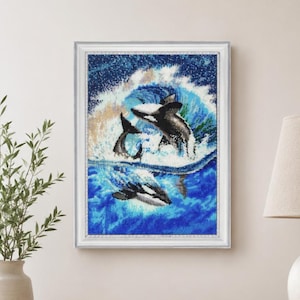 May include: A framed diamond painting featuring a black and white orca whale breaching through a vibrant blue ocean wave, with a smaller orca visible below the surface, creating a dynamic scene of marine life rendered in sparkling bead textures.
