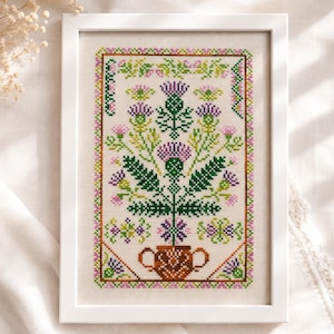 Peut inclure: Une broderie au point de croix encadrée représentant un chardon central dans des tons de vert et de violet, placé dans un pot marron à deux anses. Le motif est entouré d'une bordure géométrique décorative en points roses, verts et violets.