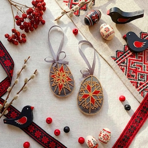 May include: An Easter-themed display featuring two gray, egg-shaped ornaments with red and gold starburst designs. Wooden black birds with red accents and decorated eggs are scattered around. Red berries and a patterned ribbon add color.