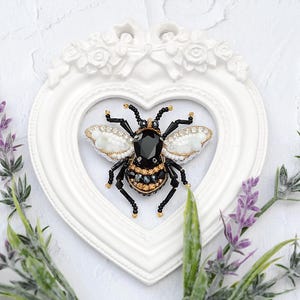 Può includere: Una spilla a forma di ape con perline, caratterizzata da una grande pietra nera sfaccettata centrale, ali ricamate con perline bianche e dorate e zampe con perline nere, posizionata all'interno di una cornice bianca a forma di cuore con rilievi floreali.