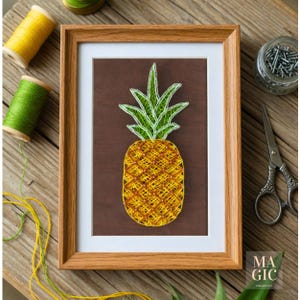 Op de afbeelding: Een ingelijst ananas draadkunstwerk. De ananas is gemaakt met gele en oranje draad op een bruine achtergrond, met groene draad voor de bladeren. Het kunstwerk zit in een houten lijst, met spoelen garen en een schaar in de buurt.
