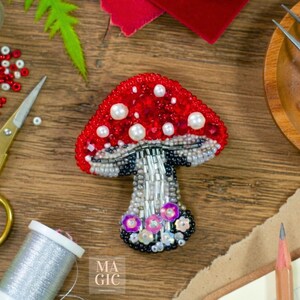 May include: A handcrafted mushroom brooch with a red beaded cap and white pearl accents. The stem is silver and black, with a cluster of pink and purple flowers at the base. The brooch is embellished with sequins and beads.