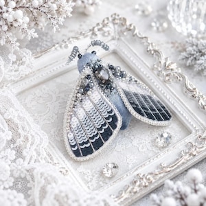 May include: A moth-shaped brooch featuring a gradient of white to black sequins on its wings. The body is crafted from light blue fabric, accented with dark grey beads and a central faceted crystal. Two coiled wire antennae extend from the head.