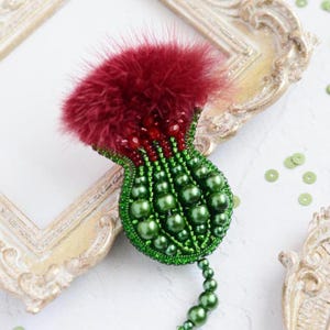 May include: A thistle-shaped brooch featuring a deep red fluffy top, a band of small red beads, and a base constructed from various sizes of glossy green pearls. The piece is intricately stitched with green thread, creating a textured, rounded form.