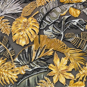 May include: Detailed textile featuring embroidered tropical leaves in gold, black, and white. The leaves are embellished with beadwork, set against a dark grey fabric background. The design showcases a variety of leaf shapes.