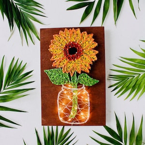 Pode incluir: Arte de fio de girassol num frasco de pedreiro com luzes de fadas num fundo de madeira. O girassol é feito com fio laranja e amarelo, as folhas são verdes e o frasco é delineado com fio branco.