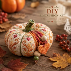 May include: A fabric pumpkin pincushion featuring cross-stitch floral patterns in orange, yellow, and red. It includes a green ceramic stem, yarn tassels, wooden beads, and an orange heart-shaped charm. Text reads: DIY PINCUSHION KIT.