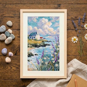 May include: Framed cross-stitch art showing a coastal scene with a white house, blue roof, and red chimney. The artwork features a beach, ocean waves, and lavender and white flowers. The frame is light-coloured wood.