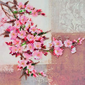 Op de afbeelding: Een kralen bloemkunstwerk met roze kersenbloesems met witte en gouden centra en bruine stelen. De bloesems zijn gerangschikt op een canvas met een zachte, patroonachtergrond in roze en beige tinten.