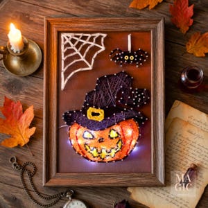 Peut inclure: Une œuvre d'art en ficelle d'Halloween encadrée, représentant une citrouille-lanterne lumineuse avec un chapeau de sorcière, une araignée et une toile. Le cadre est marron et le fond est brun foncé.