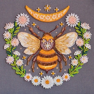 May include: A beaded artwork featuring a bee with detailed wings and a striped abdomen, surrounded by a floral wreath. A crescent moon and stars are above the bee. The color palette includes gold, brown, white, and green beads on a gray background.