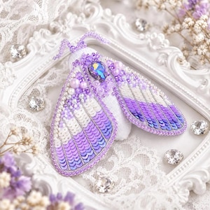 May include: A moth-shaped brooch featuring purple and white sequins, small white pearls, and a central iridescent teardrop-shaped crystal. The wings are detailed with layered rows of sequins, and the body is adorned with beads and antennae.