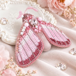 May include: A moth-shaped brooch crafted with pink and burgundy sequins, beads, and crystals. The wings feature a gradient pattern of light pink to dark burgundy sequins, accented with white pearls and a central clear crystal on the body.