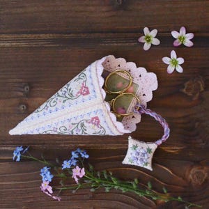 May include: A cone-shaped fabric holder featuring intricate floral cross-stitch embroidery in shades of pink, blue, and green, finished with a pale pink scalloped lace trim. Inside are gold-toned metal scissors attached to a small square pincushion.