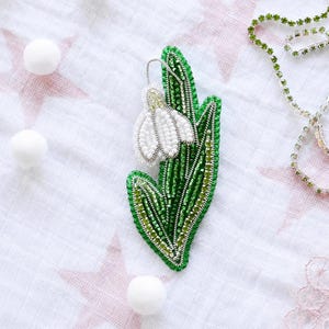 Peut inclure: Une broche perlée faite à la main en forme de perce-neige. Le motif présente une fleur blanche perlée avec des accents de fil métallique argenté, contrastant avec des feuilles perlées aux nuances variées de vert émeraude et vert lime.