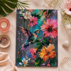 Op de afbeelding: Een diamond painting set met een kolibrie in vlucht naar een grote roze hibiscusbloem. Het kunstwerk bevat levendige oranje en roze bloesems, groene bladeren en een donkergroenblauwe achtergrond, allemaal gemaakt met fonkelende kralen.