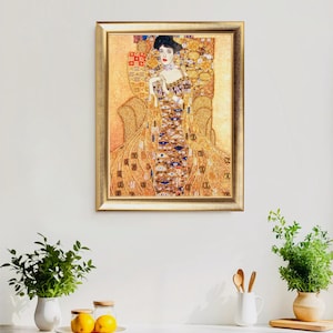 K&ouml;nnte beinhalten: Ein gerahmter Druck eines Gem&auml;ldes von Gustav Klimt. Das Kunstwerk zeigt eine Frau in einem aufwendigen Kleid, die in einem Stuhl sitzt. Die Farbpalette umfasst Gold-, Braun- und Blaut&ouml;ne. Der Rahmen ist goldfarben. Das Kunstwerk wird an einer wei&szlig;en Wand ausgestellt.
