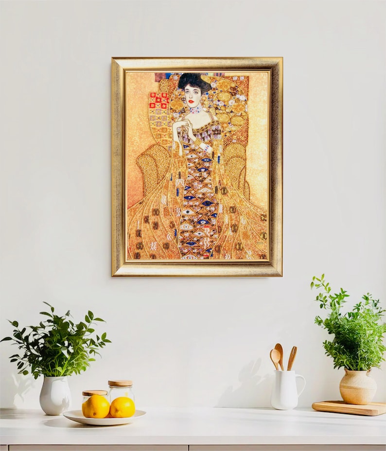 K&ouml;nnte beinhalten: Ein gerahmter Druck eines Gem&auml;ldes von Gustav Klimt. Das Kunstwerk zeigt eine Frau in einem aufwendigen Kleid, die in einem Stuhl sitzt. Die Farbpalette umfasst Gold-, Braun- und Blaut&ouml;ne. Der Rahmen ist goldfarben. Das Kunstwerk wird an einer wei&szlig;en Wand ausgestellt.