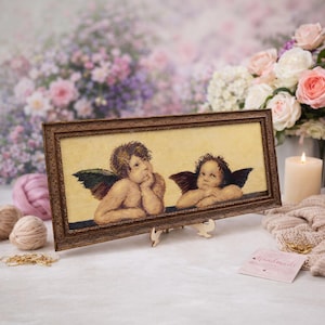 May include: A framed cross-stitch artwork featuring two cherubs with detailed wings and expressions. The artwork is set against a light yellow background and framed in a dark brown, ornate frame. The piece is displayed on a wooden easel.