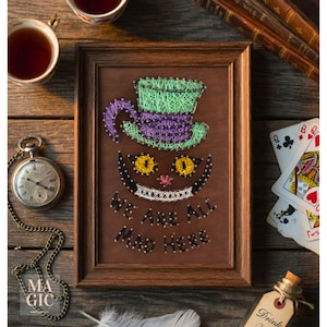 Könnte beinhalten: Ein gerahmtes String-Art-Stück mit der Grinsekatze aus Alice im Wunderland. Das Gesicht und der Hut der Katze sind mit farbigem Faden auf braunem Hintergrund gefertigt. Der Text "WE ARE ALL MAD HERE" befindet sich unter dem Grinsen der Katze. Eine Taschenuhr und Spielkarten sind in der Nähe.