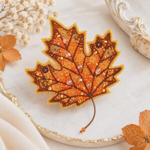 Peut inclure: Une broche en forme de feuille d'érable ornée de perles de rocaille orange et marron, de sequins et de cristaux à facettes. La feuille présente une structure nervurée détaillée soulignée par des perles marron foncé et une tige fine.
