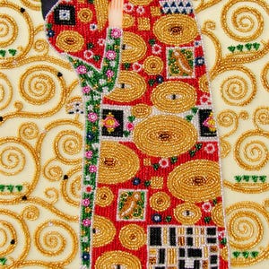 K&ouml;nnte beinhalten: Nahaufnahme eines dekorativen Kunstwerks mit einer Figur in einem roten Gewand, das mit goldenen Verzierungen geschm&uuml;ckt ist. Das Gewand hat ein Muster aus goldenen Kreisen und Quadraten. Der Hintergrund ist cremefarben mit goldenen Wirbeln.