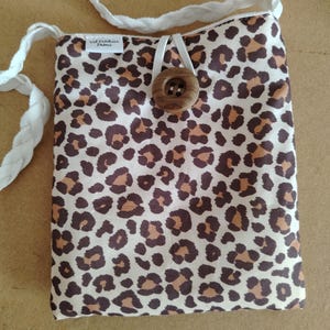 Peut inclure: Un sac en tissu rectangulaire avec un motif léopard marron et noir sur fond crème. Le sac comprend une bandoulière tressée blanche, une grande fermeture à bouton en bois marron et une petite étiquette blanche portant l'inscription Val Créations Drôme.