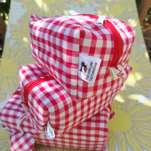 Peut inclure: Une pile de trois trousses rectangulaires en tissu à motif vichy rouge et blanc. Chaque trousse possède une fermeture éclair rouge et une petite étiquette en tissu blanc avec le texte Fait main par Val Créations et une icône de machine à coudre.