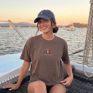 Könnte beinhalten: Braunes T-Shirt mit einer kleinen Grafik eines Fuchses, der eine Kaffeetasse hält, und dem Text "Main Character Energy". Die Person trägt eine marineblaue Kappe und goldene Ohrringe. Der Hintergrund zeigt einen Sonnenuntergang über dem Wasser.