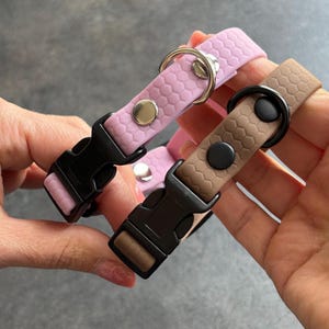 Può includere: Vengono mostrati due collari per cani, uno rosa chiaro e uno marrone. Ogni collare presenta una fibbia nera, un anello a D in metallo e un motivo a nido d'ape strutturato. I collari sono tenuti su uno sfondo grigio.