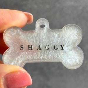 Op de afbeelding: Een botvormig huisdierlabel met de naam "SHAGGY" in zilveren letters. Het label heeft een parelmoer, glinsterende witte afwerking en een gat om aan een halsband te bevestigen. Een huisdieraccessoire.