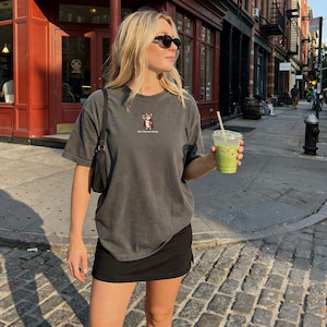 Peut inclure: T-shirt gris foncé avec un petit motif et le texte "More Chaotic Energy". La tenue comprend une jupe noire et des lunettes de soleil. La personne tient une boisson verte dans un gobelet en plastique transparent avec une paille.