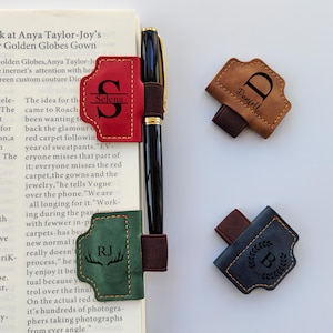 Op de afbeelding: Een zwarte pen met gouden accenten wordt getoond met verschillende leren penhouders. De penhouders zijn rood, bruin, groen en marineblauw. Elke houder heeft een andere initiaal en naam gegraveerd, met een elastiek om aan een notitieboek of boek te bevestigen.