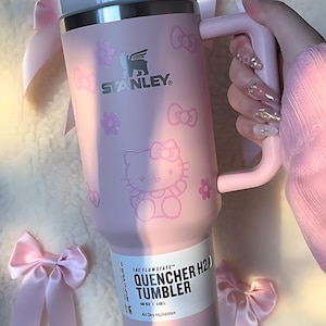 Peut inclure: Gobelet rose Stanley Quencher H2.0 avec une poignée, orné de motifs Hello Kitty et nœuds. Le gobelet de 1,18 L affiche le logo Stanley et le texte "QUENCHER H2.0 TUMBLER". Des nœuds décoratifs entourent le gobelet.