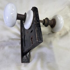 May include: A door lock mechanism featuring a rectangular black metal plate with two keyholes and three white porcelain knobs attached to a central metal spindle. The hardware displays a weathered finish with visible signs of age and use.