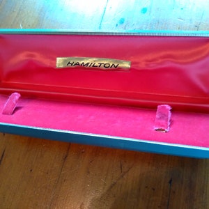 May include: An open, teal-colored box with a red velvet interior. The box has a gold label that reads "HAMILTON".